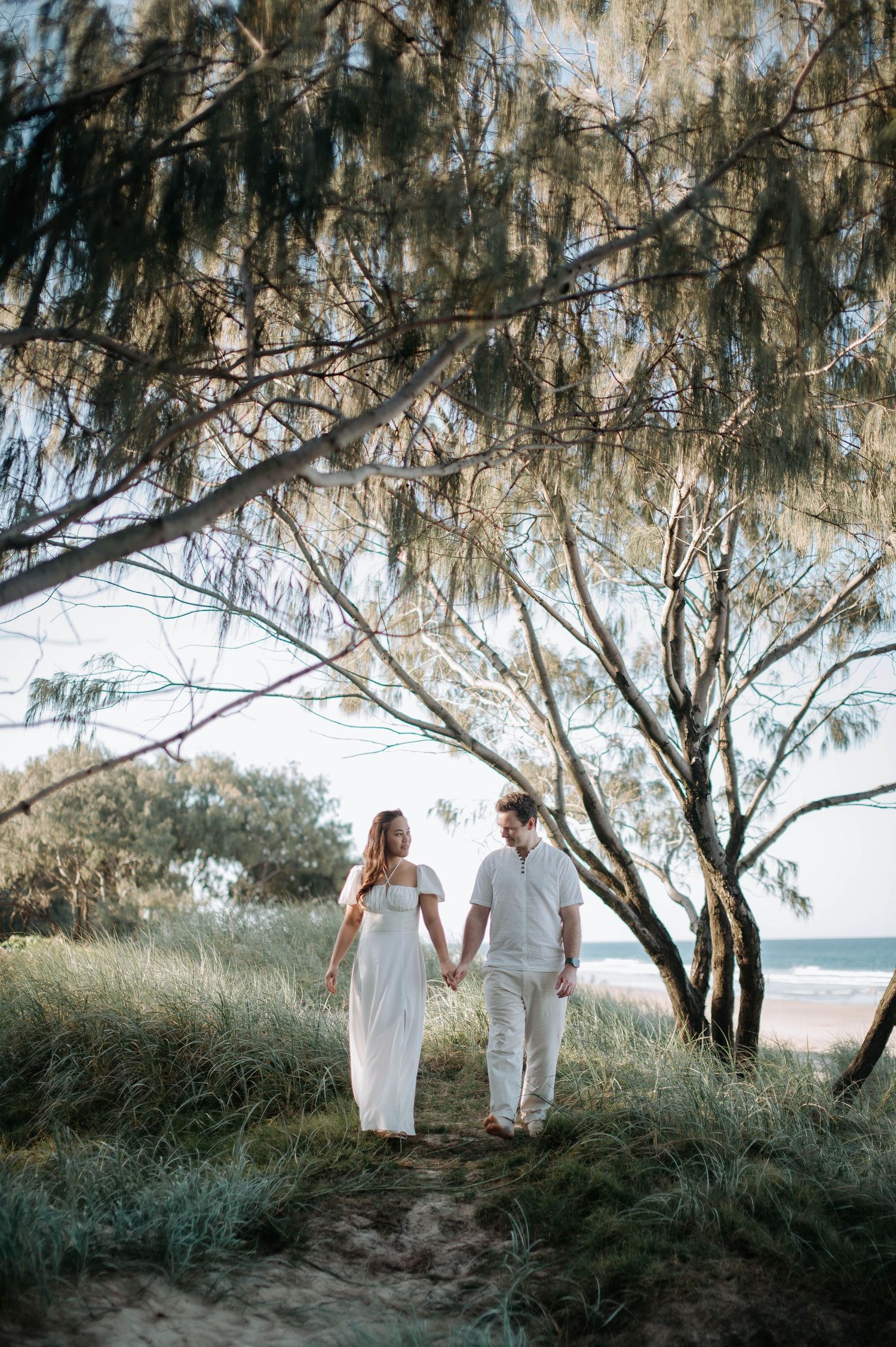Couple in white walking along a coastal path beneath overhanging beach trees.