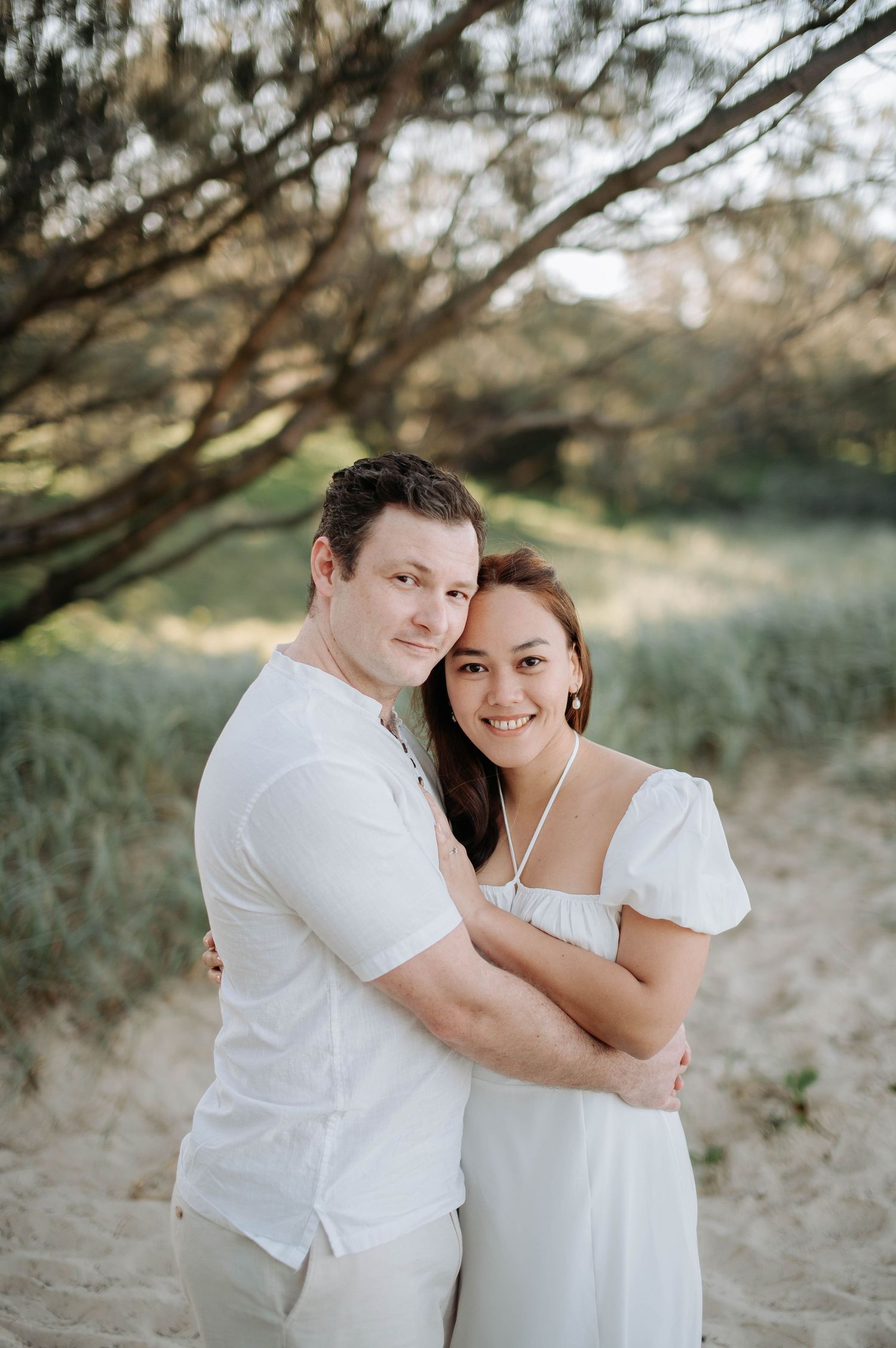 Intimate portrait of couple in white clothing embracing against nature backdrop.