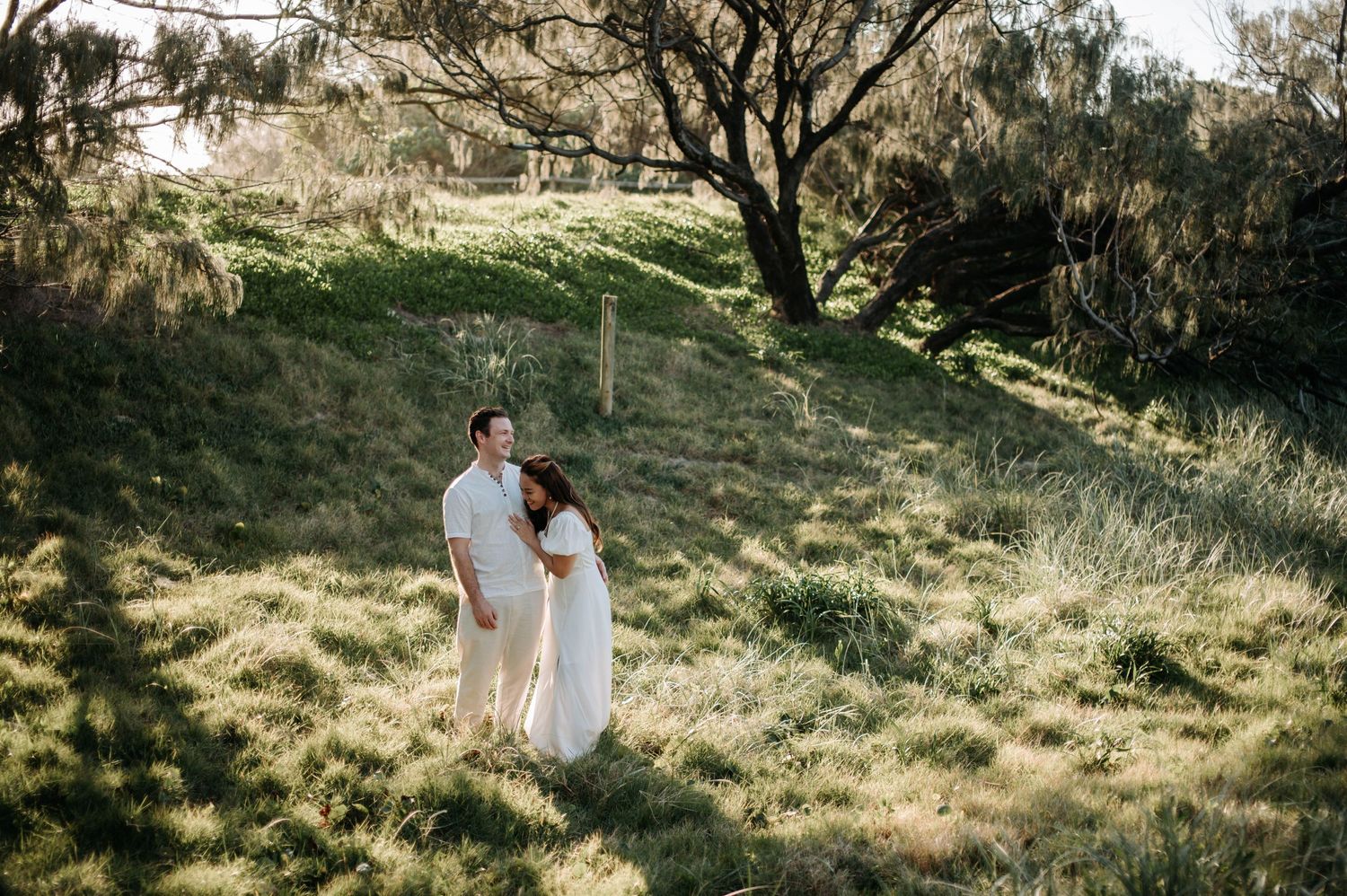 Couple in white dresswear standing in a sunlit grassy hillside setting.