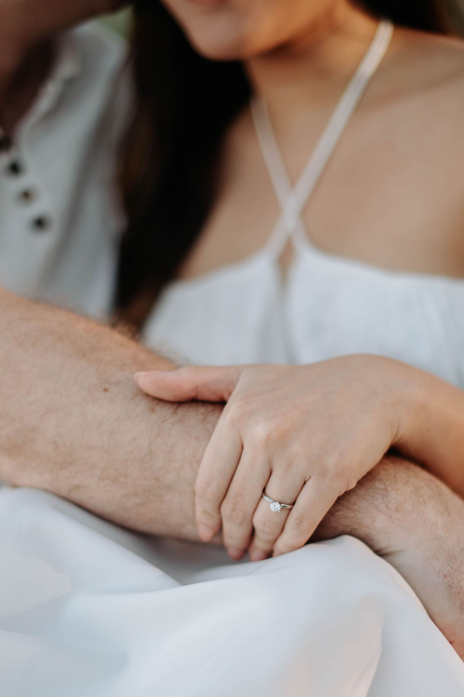 Close-up of engagement ring and hands against white clothing background.