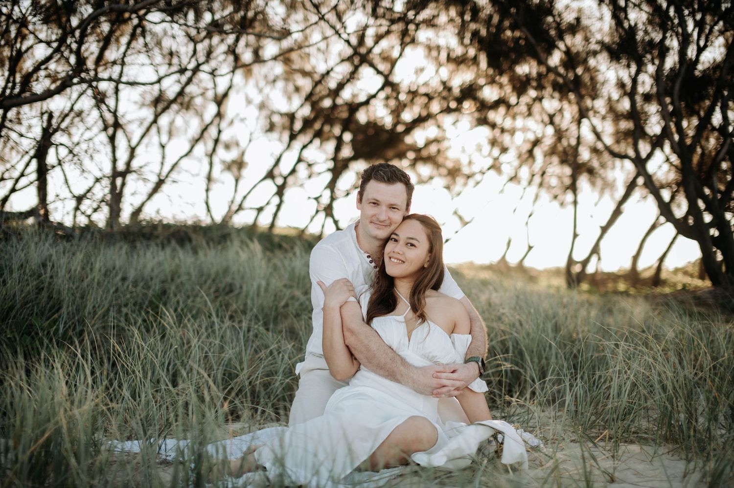 A romantic couple in white outfits embrace on a blanket during a beach photoshoot at sunset.