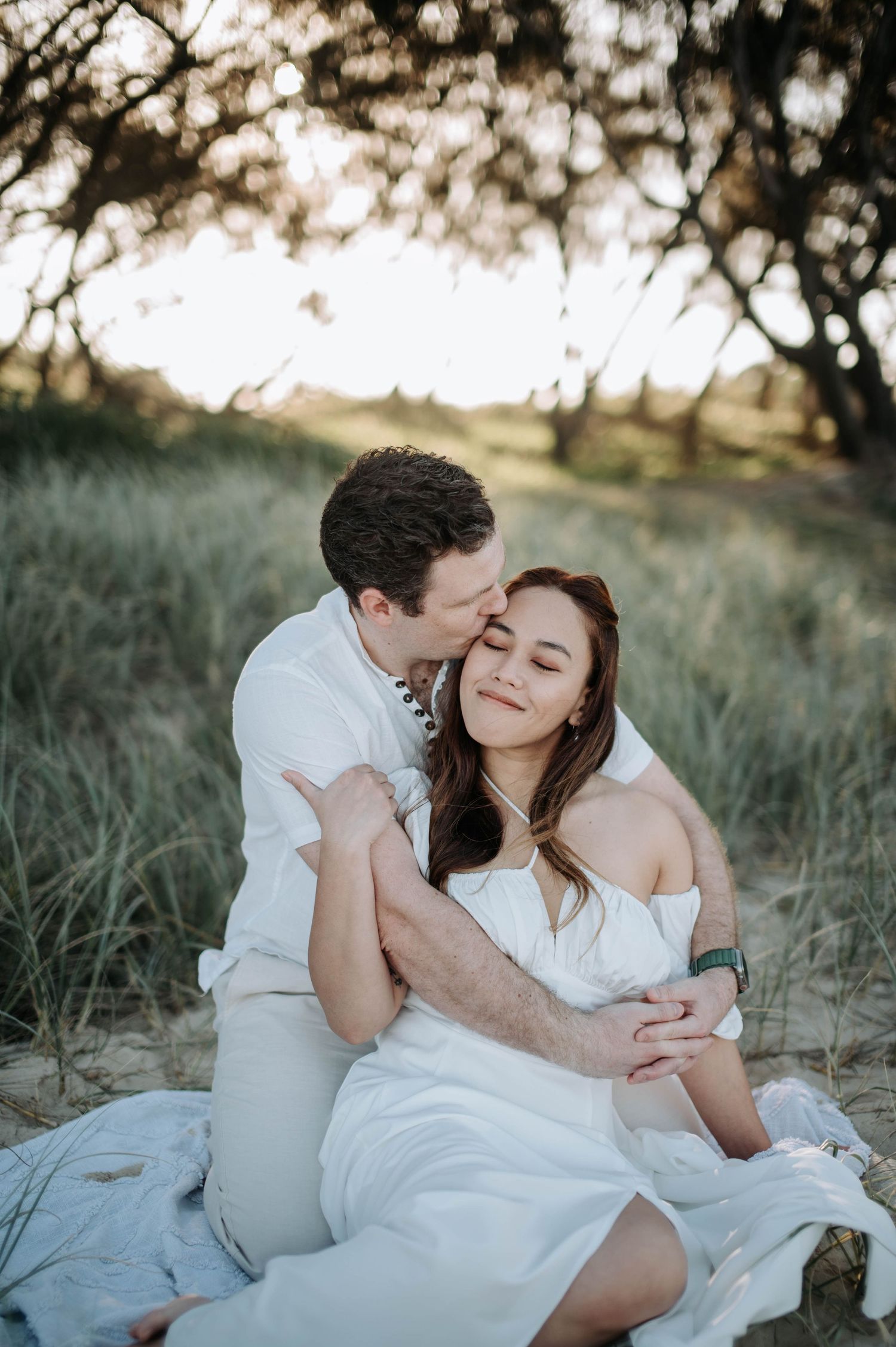 A couple shares an intimate moment while sitting on a blanket in tall grass during golden hour.