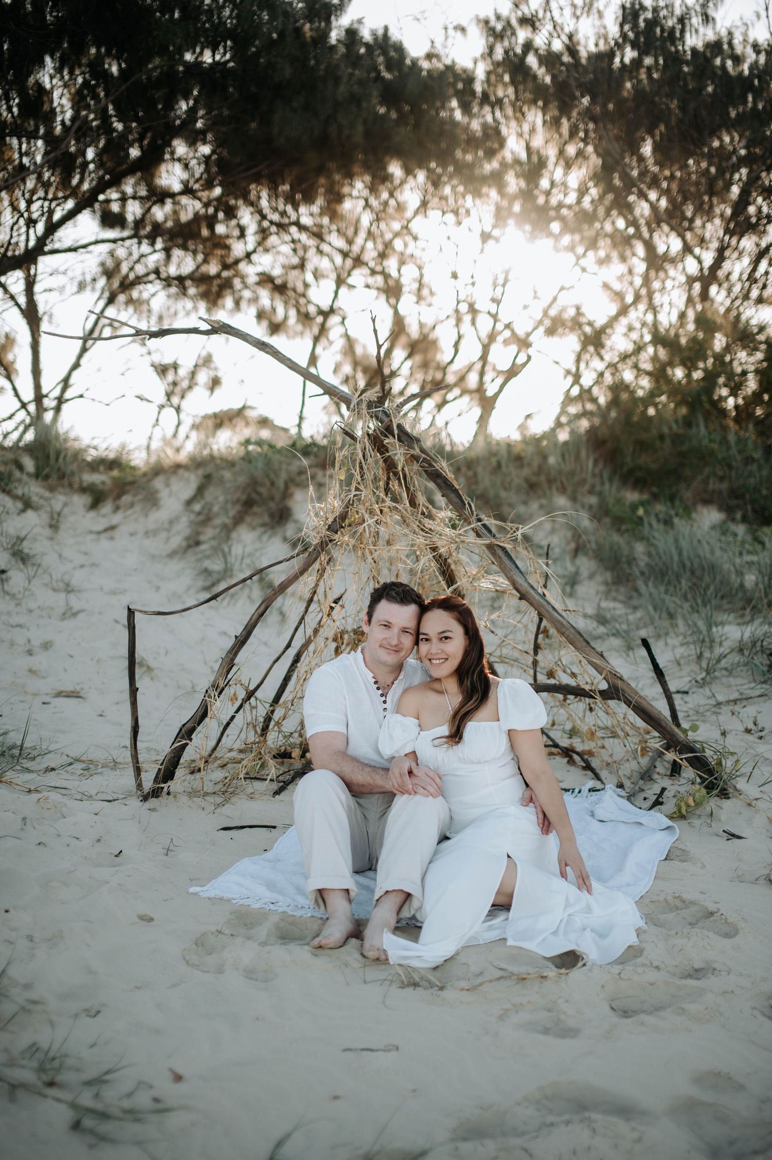 A couple poses together under a rustic driftwood teepee structure on a sandy beach.