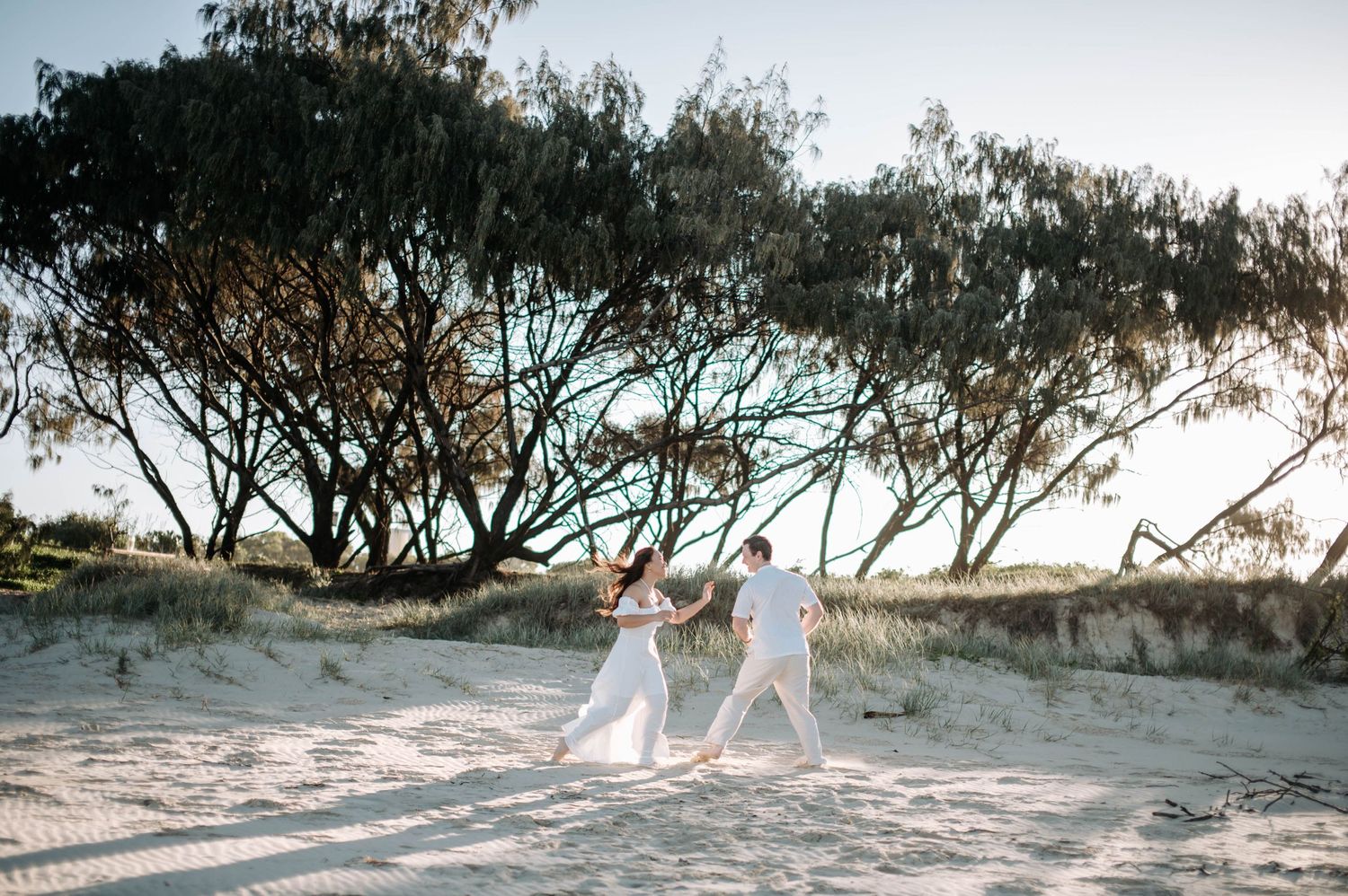 Two people in flowing white outfits walk along a coastal path with windswept trees in the background.