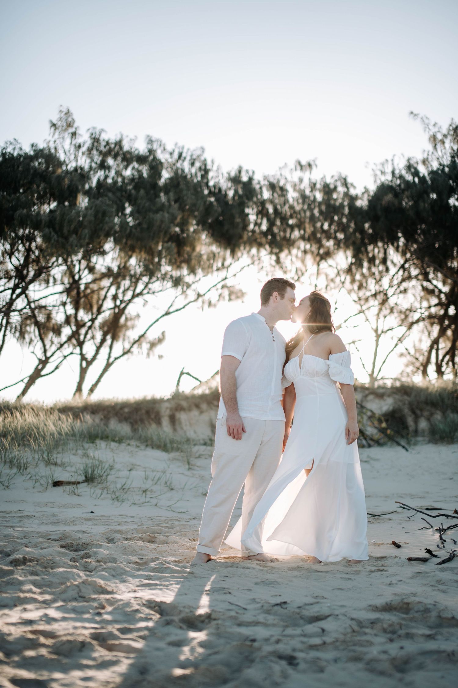 A couple in white outfits stands together on a sandy beach during sunset.