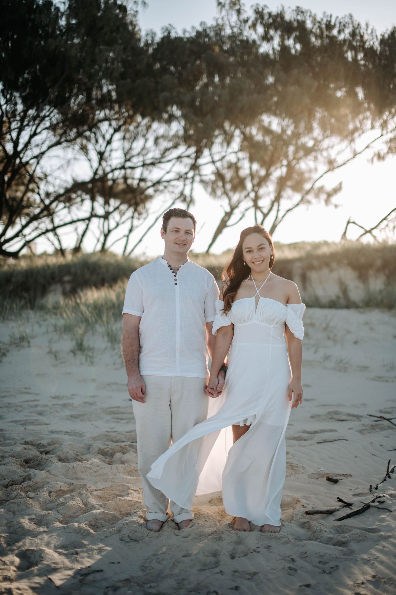 A couple poses hand-in-hand on a beach with trees silhouetted against the setting sun.