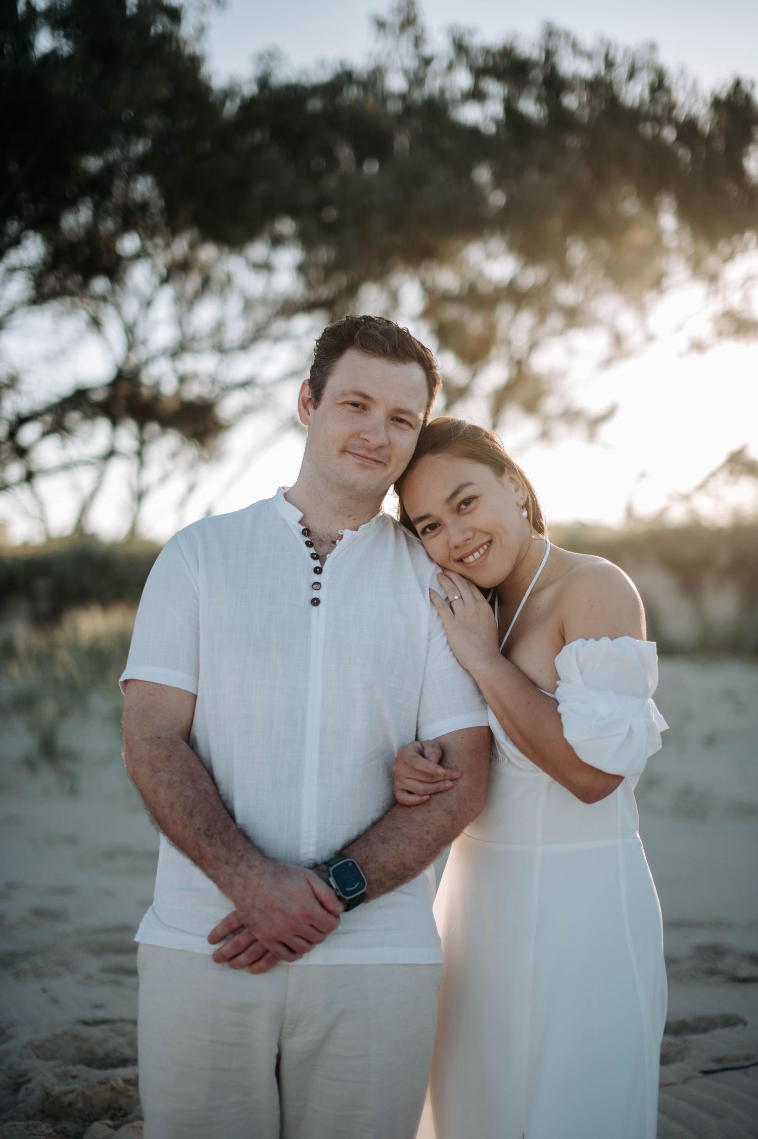 A couple in coordinating white outfits shares a tender moment during a beach sunset photoshoot.