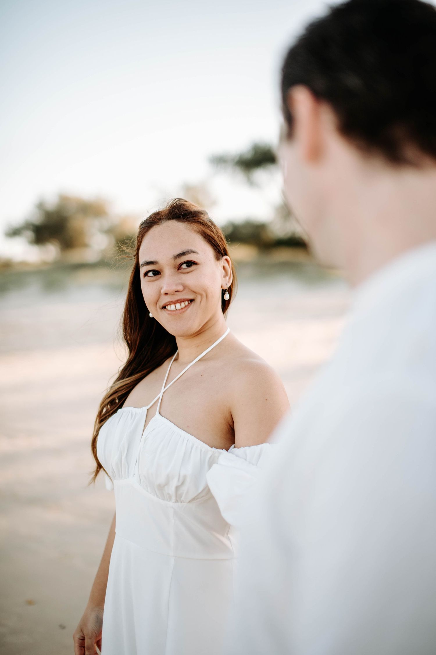A woman in a white dress smiles warmly during a romantic beach photoshoot.