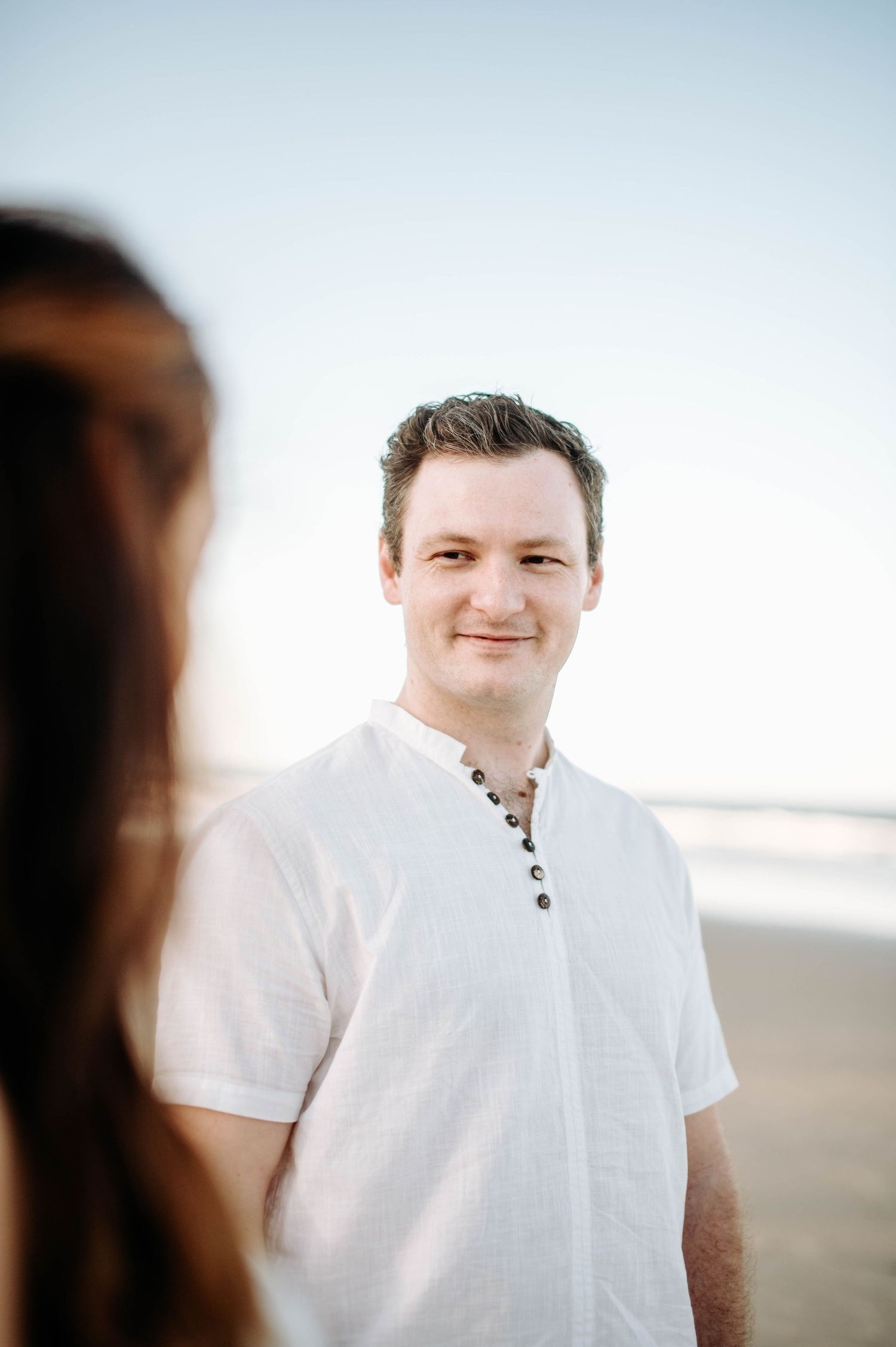 A man in a white henley shirt smiles at the beach during golden hour.