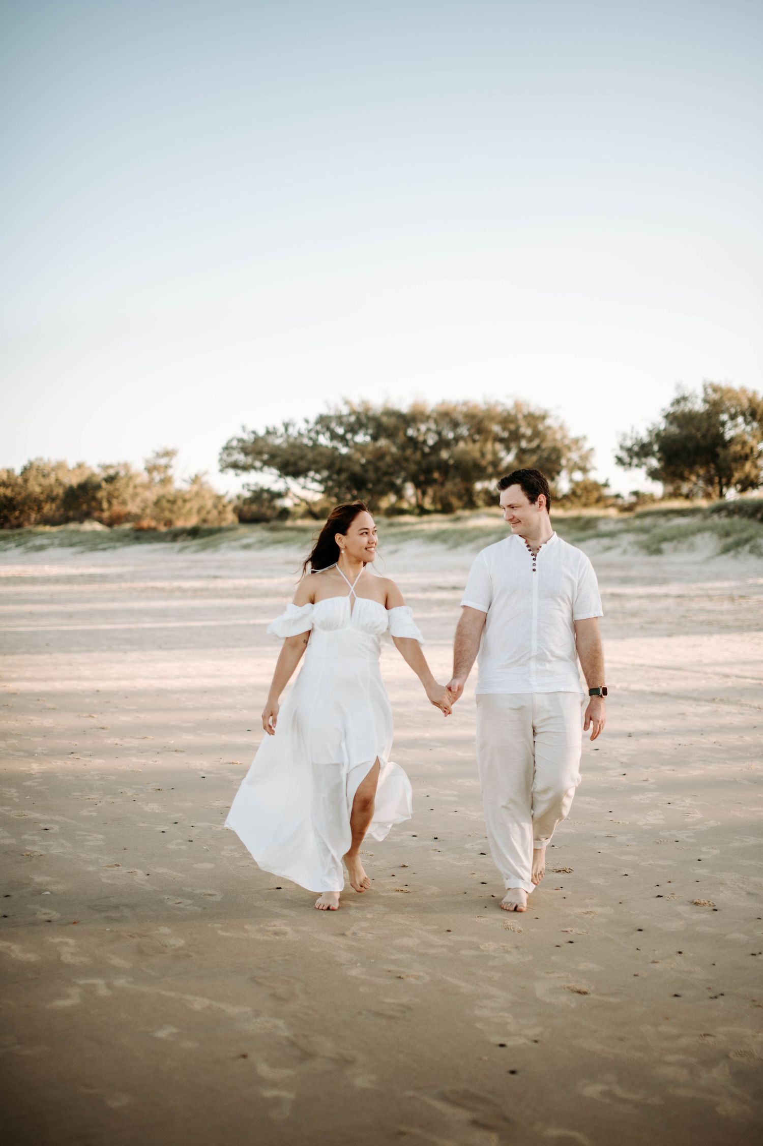 A couple walks hand in hand on a beach wearing white outfits during golden hour.