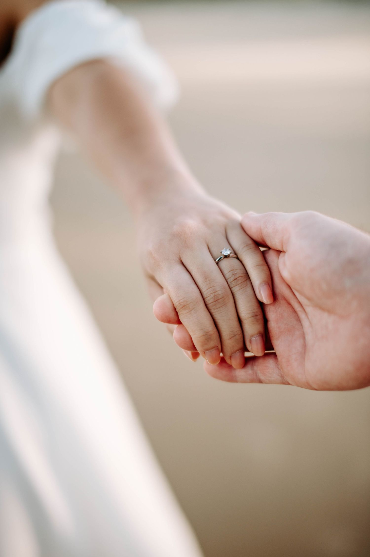 Close up of hands holding with an engagement ring visible against a blurred background.