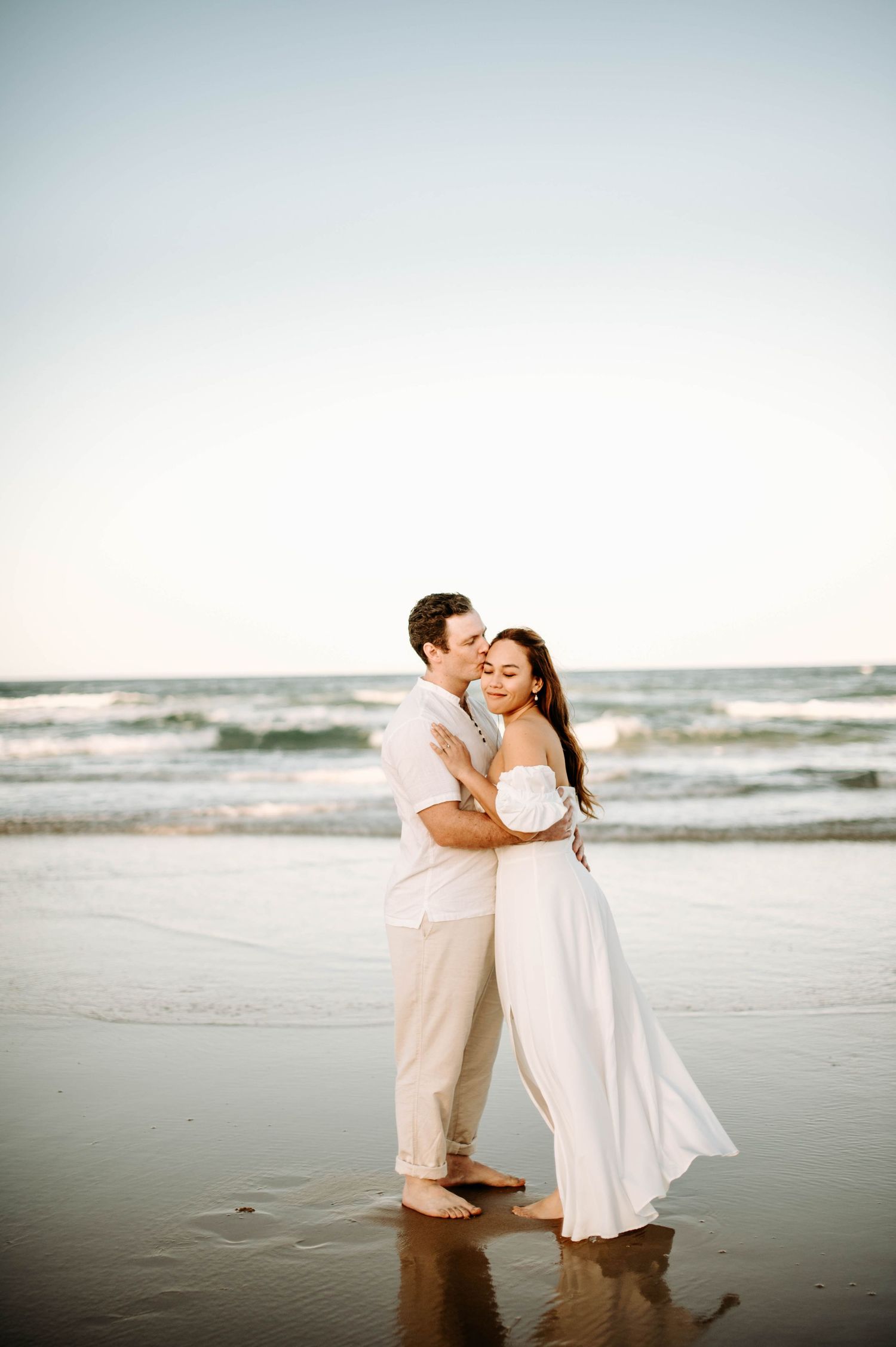 Romantic beach moment with couple in white outfits embracing by the ocean waves.