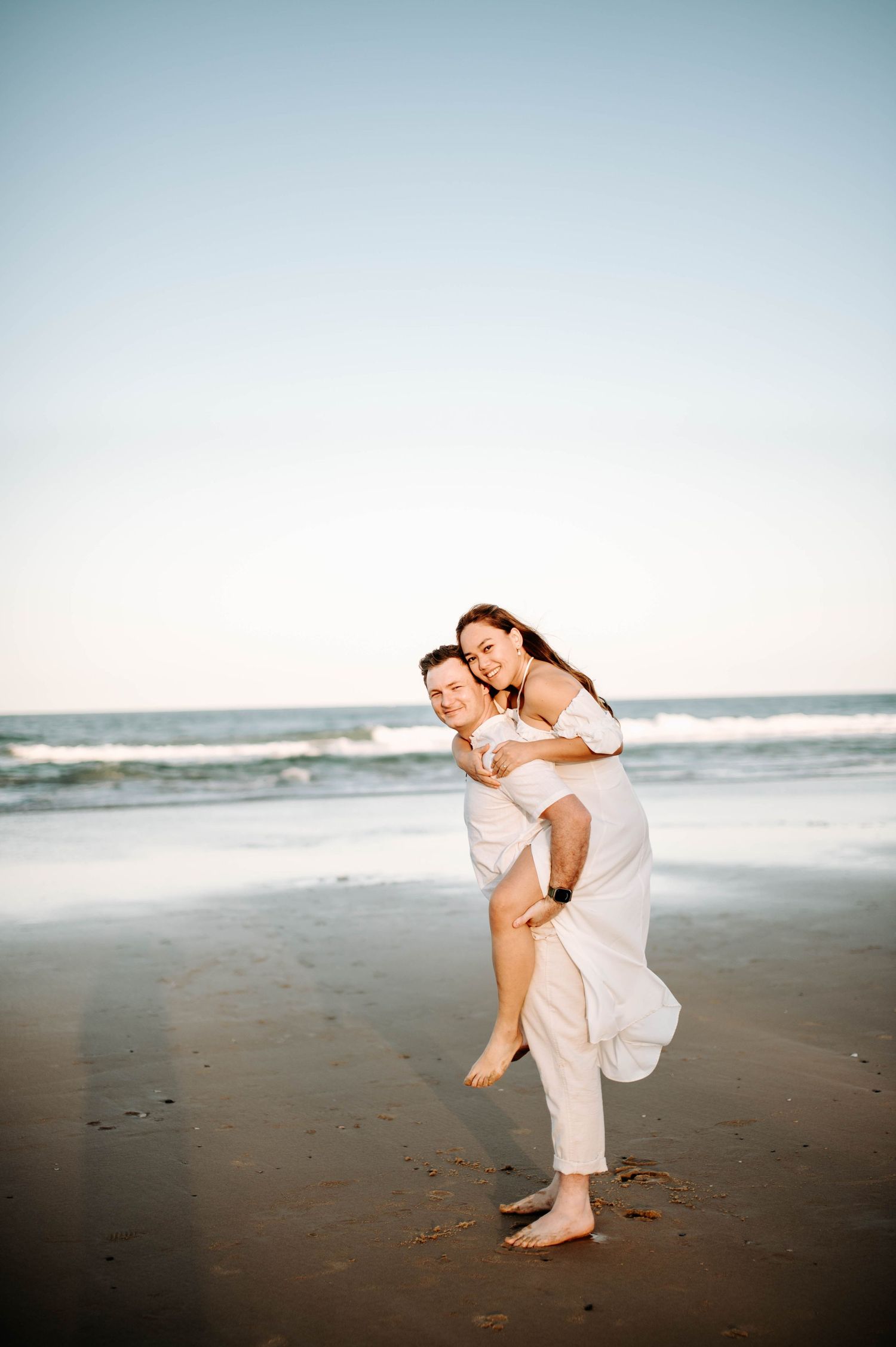 Playful beach photo with couple in white clothing having fun near ocean waves.