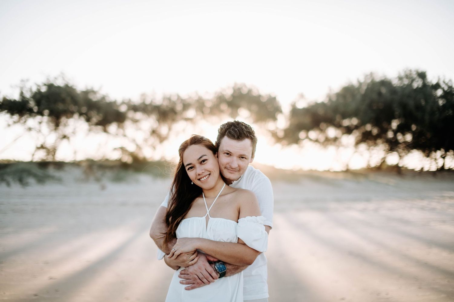 Couple embracing on beach during sunset with trees silhouetted in background.