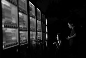 Dark silhouettes of people viewing illuminated displays in a dimly lit gallery space.