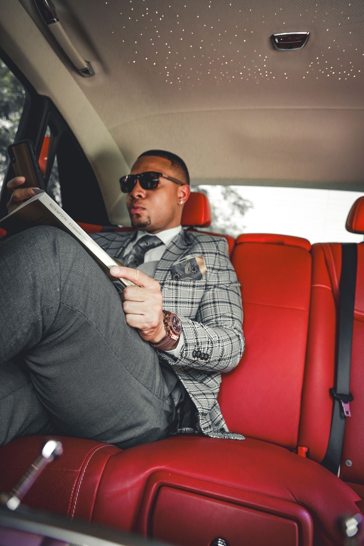 Luxury car interior showing passenger in plaid suit reading on red leather seats.