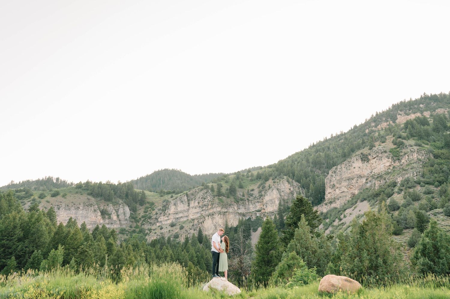 Couple standing on rocky outcrop with dramatic mountain cliffs and forest in background.