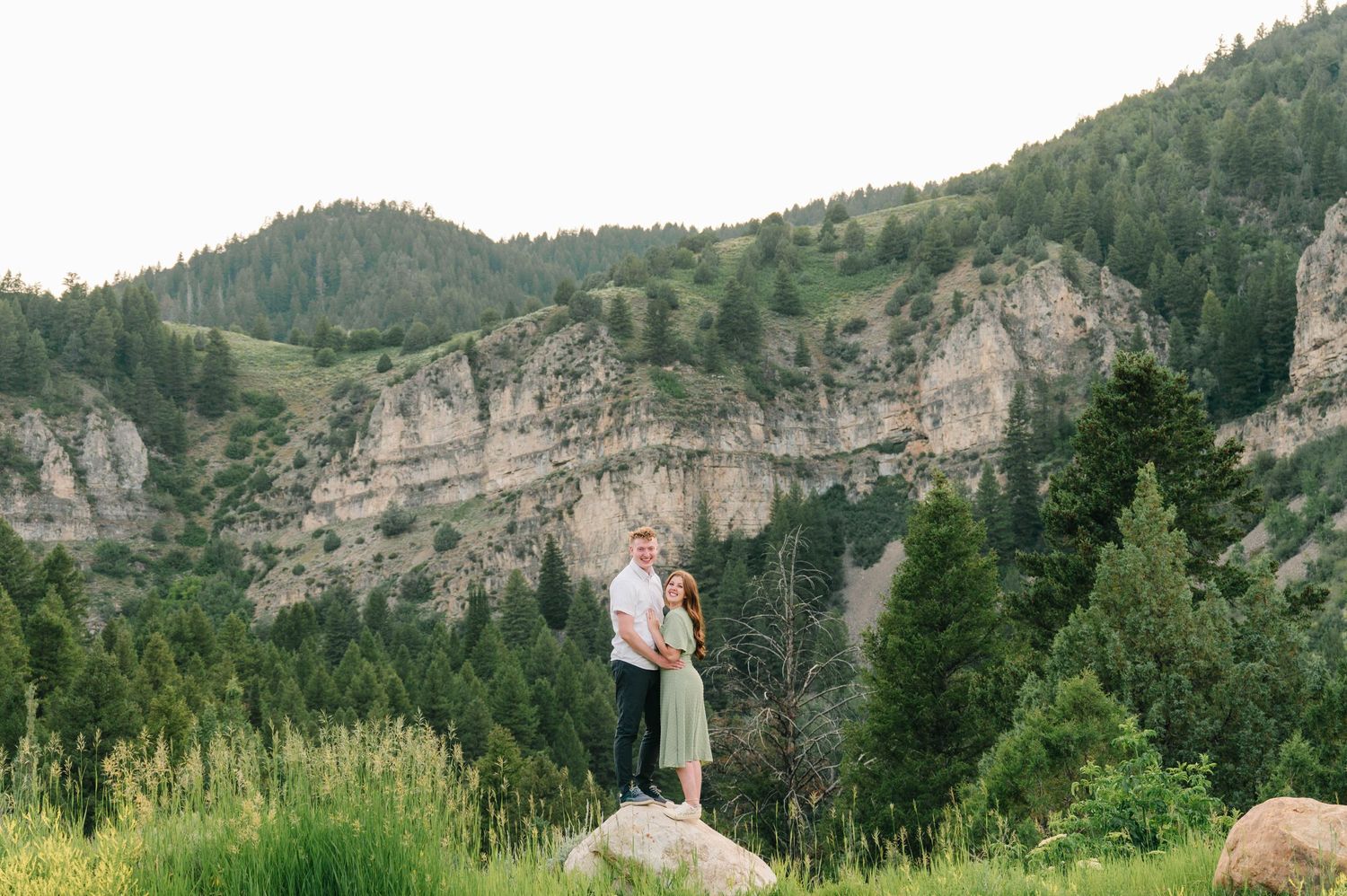 Romantic portrait of couple on boulder with majestic mountain cliffs and evergreens behind.