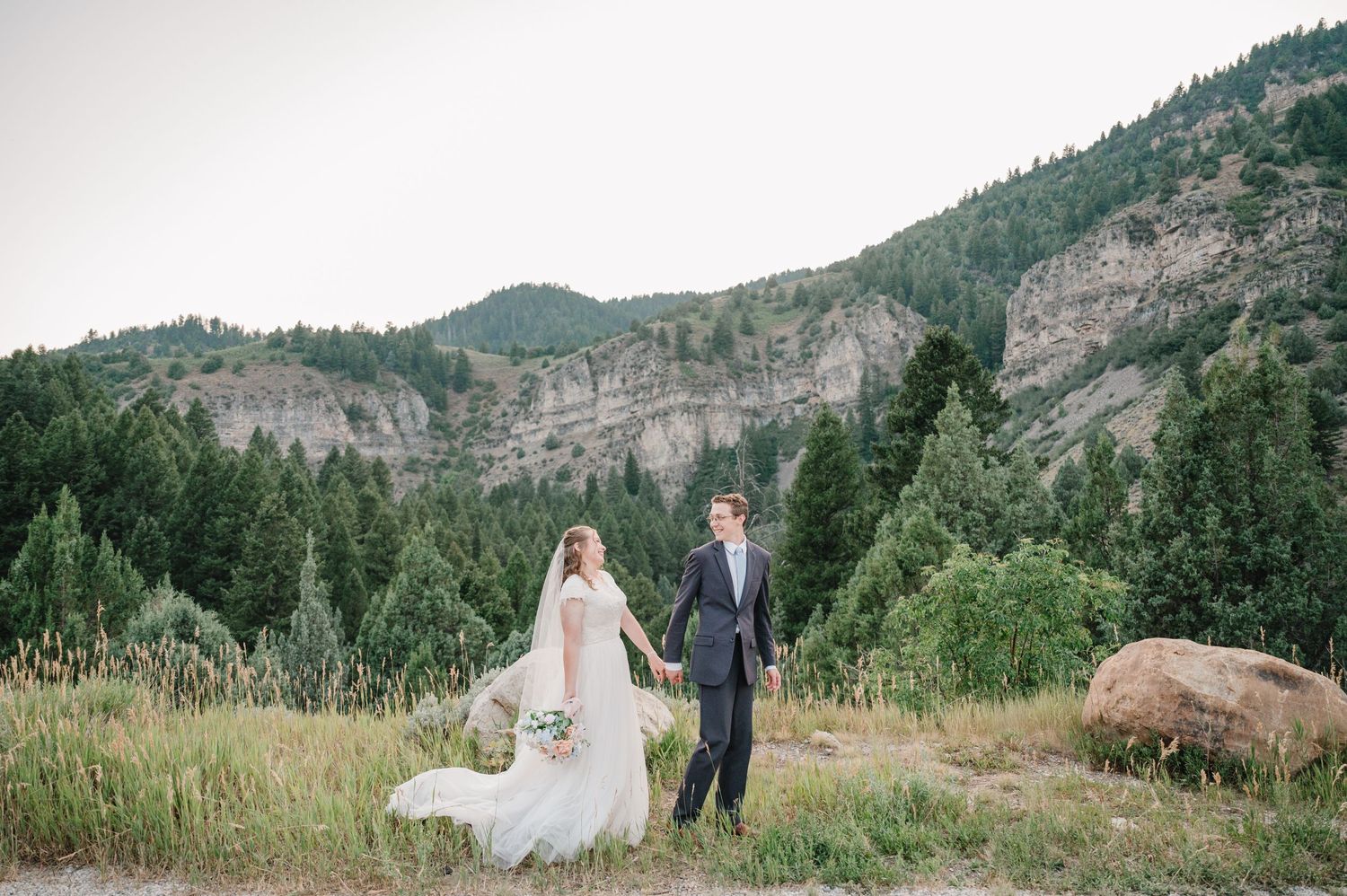 Newlyweds hold hands in mountain meadow with dramatic cliffs and evergreen forest backdrop.