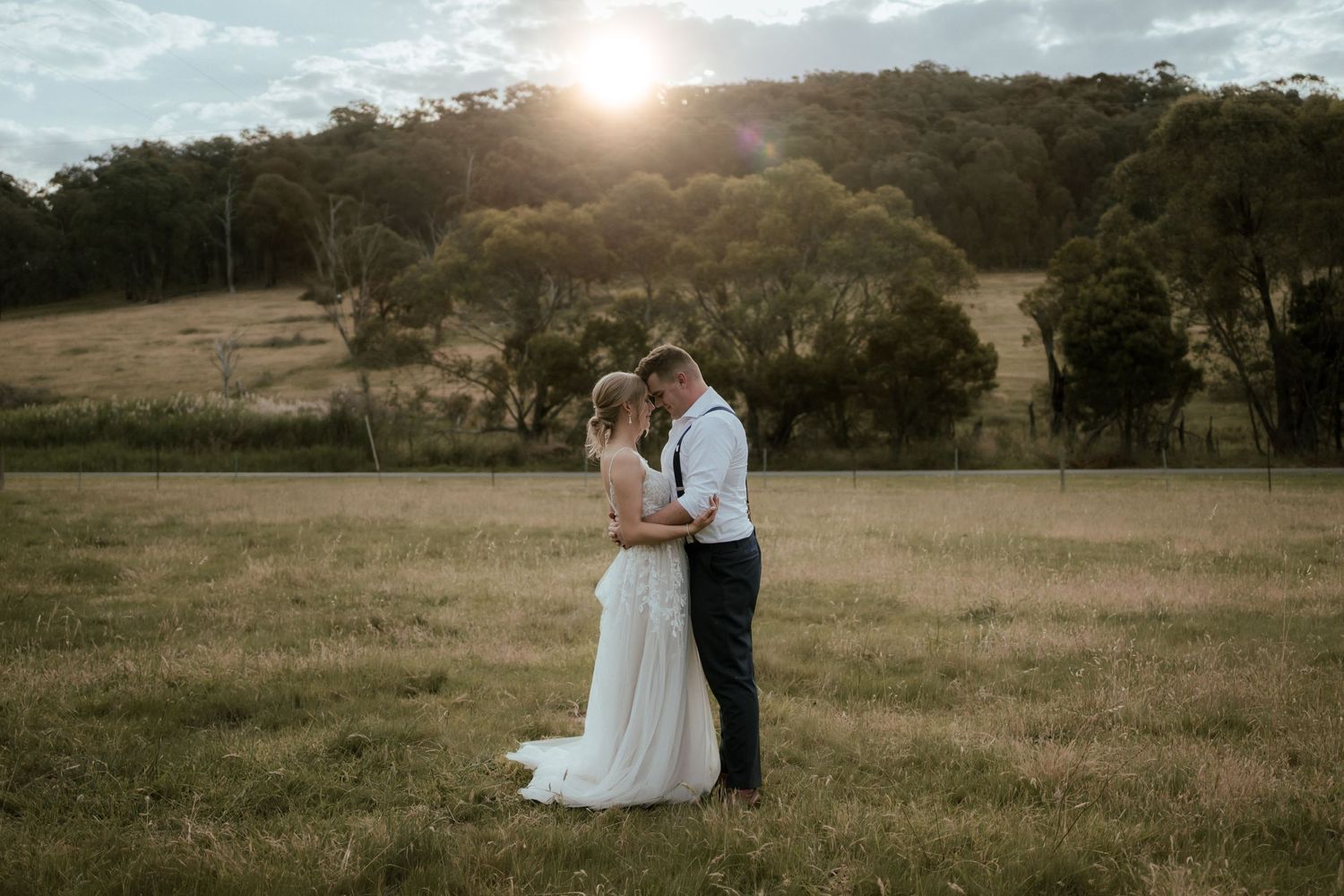 Bride and groom embrace in sunlit field with rolling hills and trees in background.