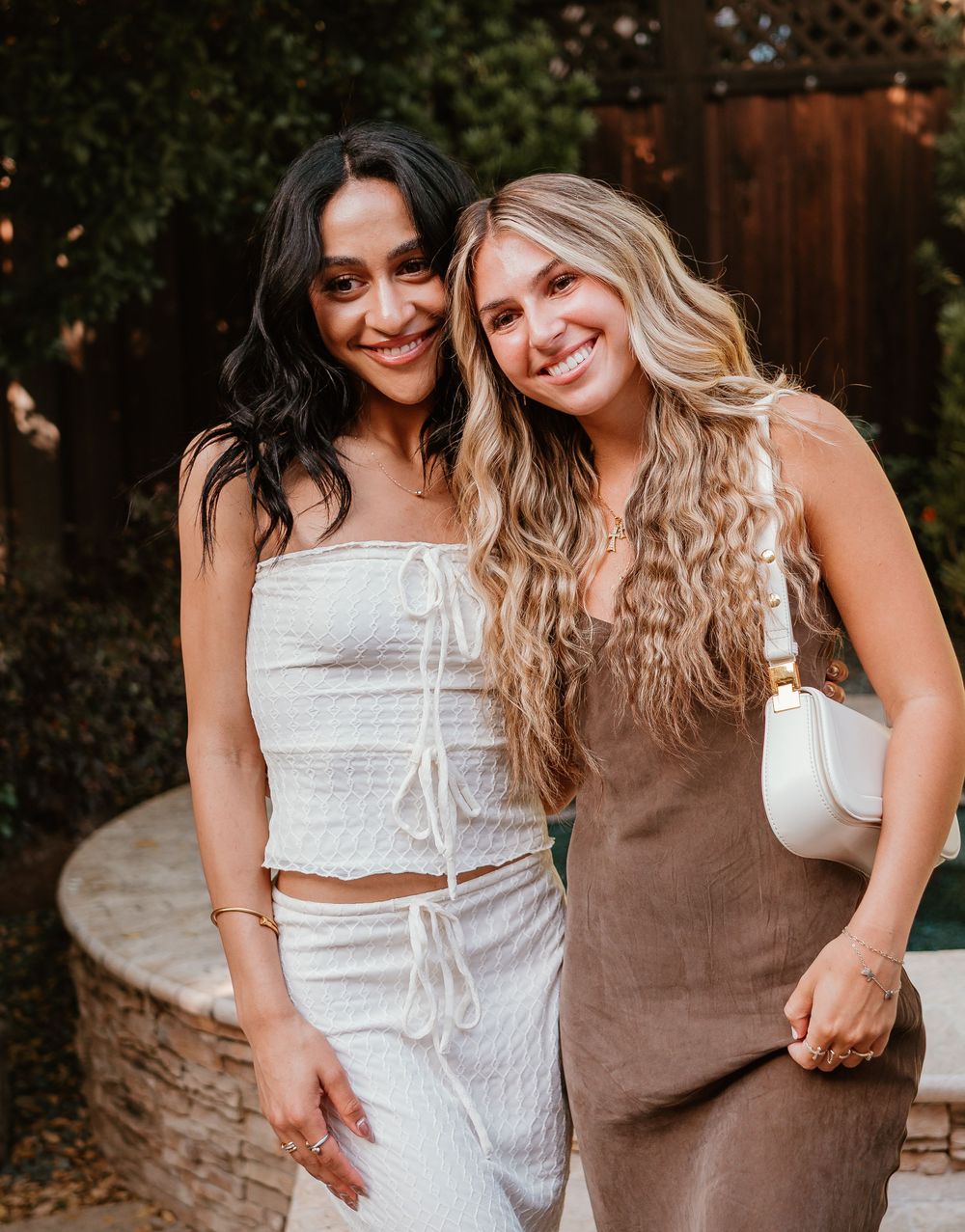 Two women in summer outfits pose together outdoors at sunset, one wearing white and one in brown.