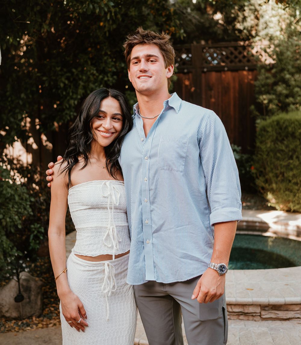A couple poses together outdoors near a pool, wearing casual summer attire in warm evening light.
