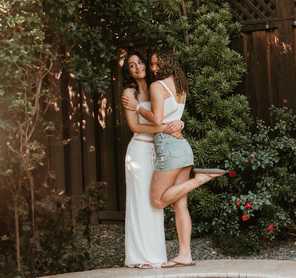 Two people in white and blue outfits embrace in an outdoor garden setting with greenery and flowers in the background.