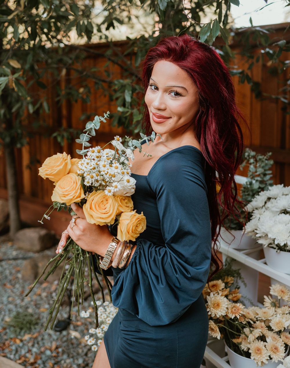 A woman in a navy dress holds a bouquet of peach roses and white flowers against a wooden fence backdrop.