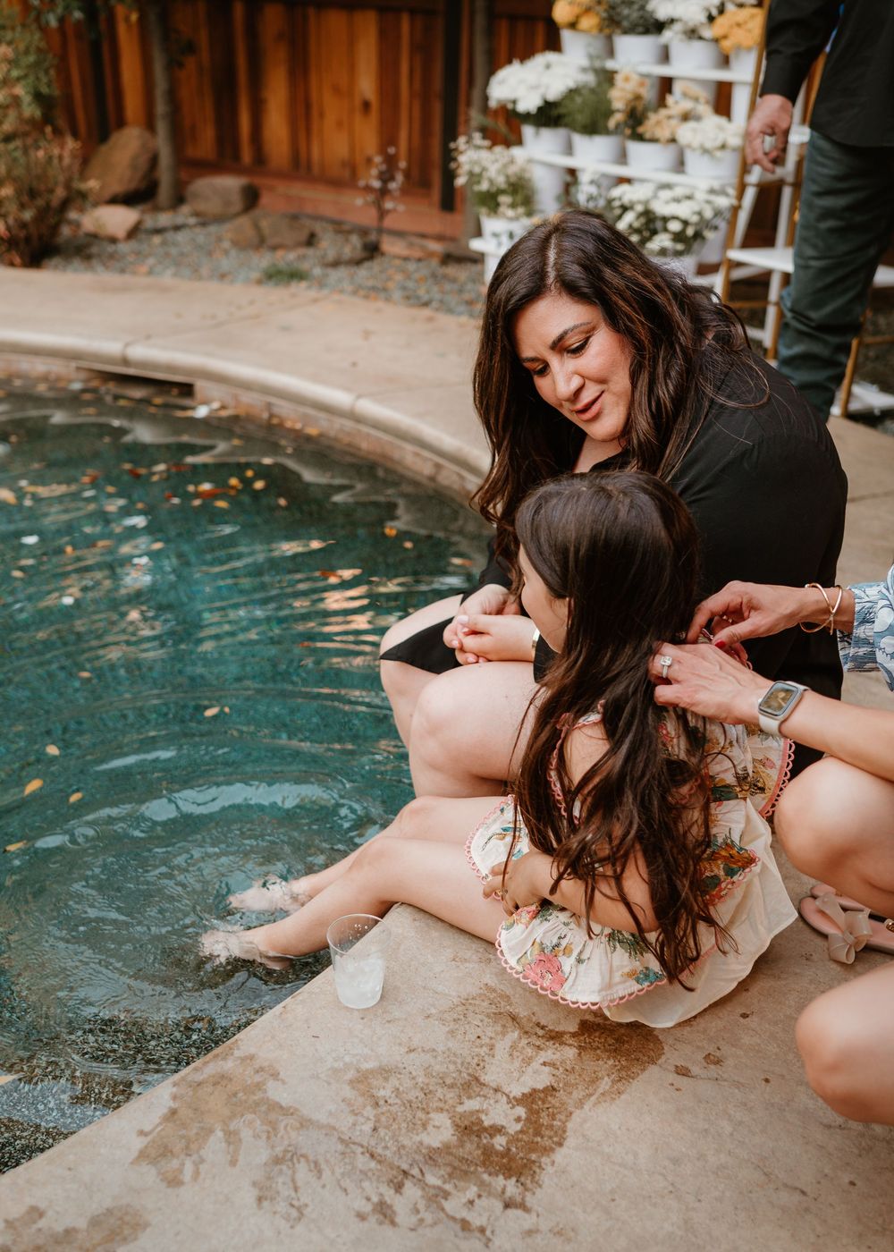 Two people sit by the edge of a pool, sharing an intimate moment in a backyard garden setting.