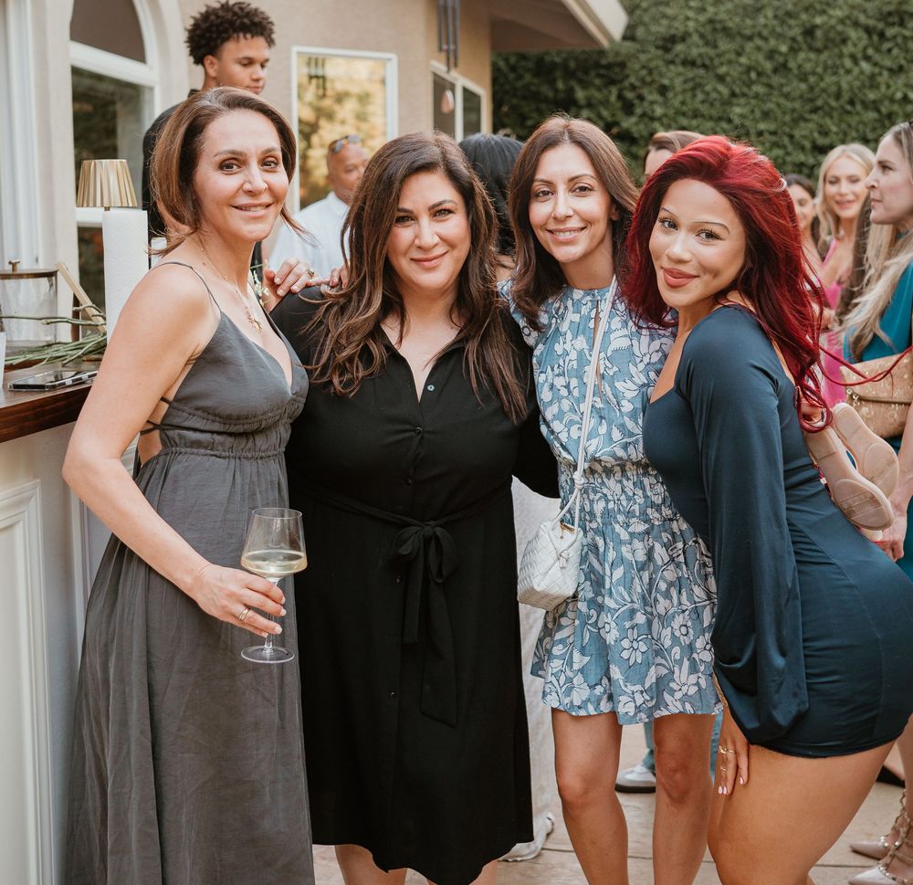 A group of friends in dresses smile together at an outdoor gathering with wine glasses and a house in the background.