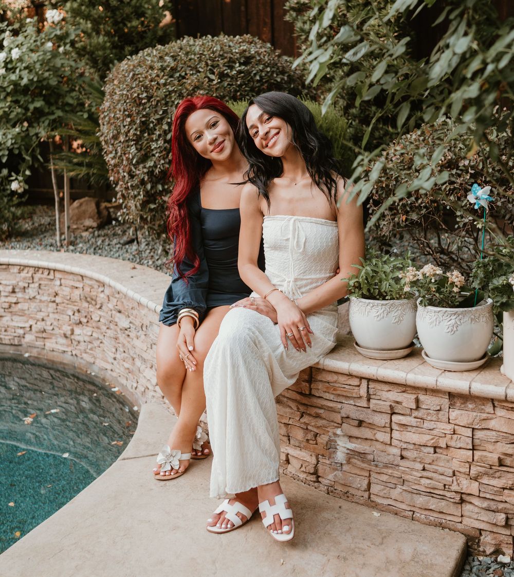 Two people in white dresses sit casually on a stone wall by a pool in a garden setting.