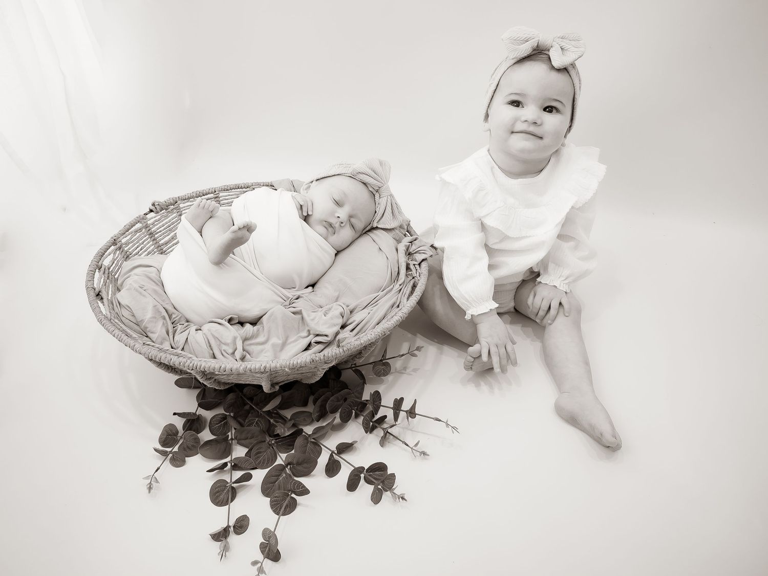 A black and white vintage-style portrait with a baby sitting next to a large basket filled with decorative flowers.