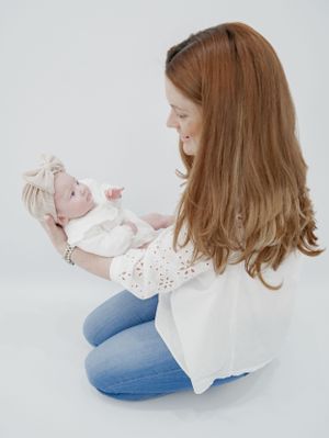 Parent kneeling on floor in white top and jeans holds small baby in white outfit against light background.