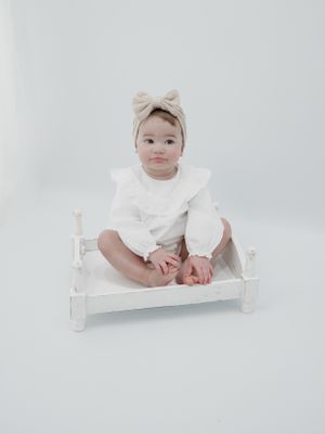 Small child wearing white outfit and beige bow headband sits on tiny white wooden bed against light background.