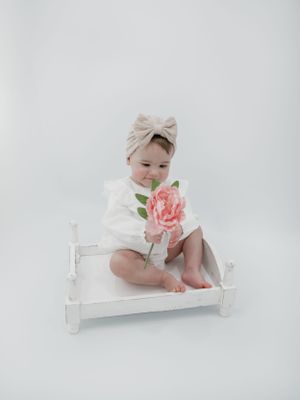 A baby wearing a white outfit and beige headband sits on a white wooden bench while holding a pink peony flower.