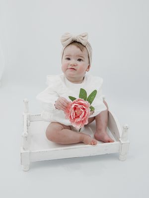 Baby in white dress sits on small white bench holding pink rose.