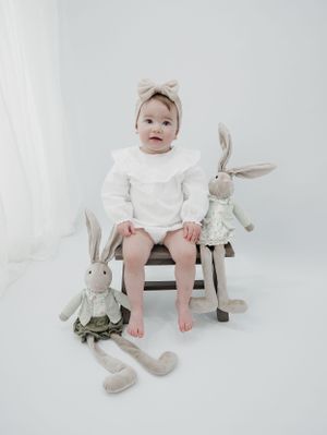 Child in white outfit sits on wooden bench with decorative bunny toys.