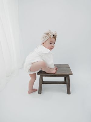 Small child in white outfit and beige headwrap climbing wooden stool against white background.
