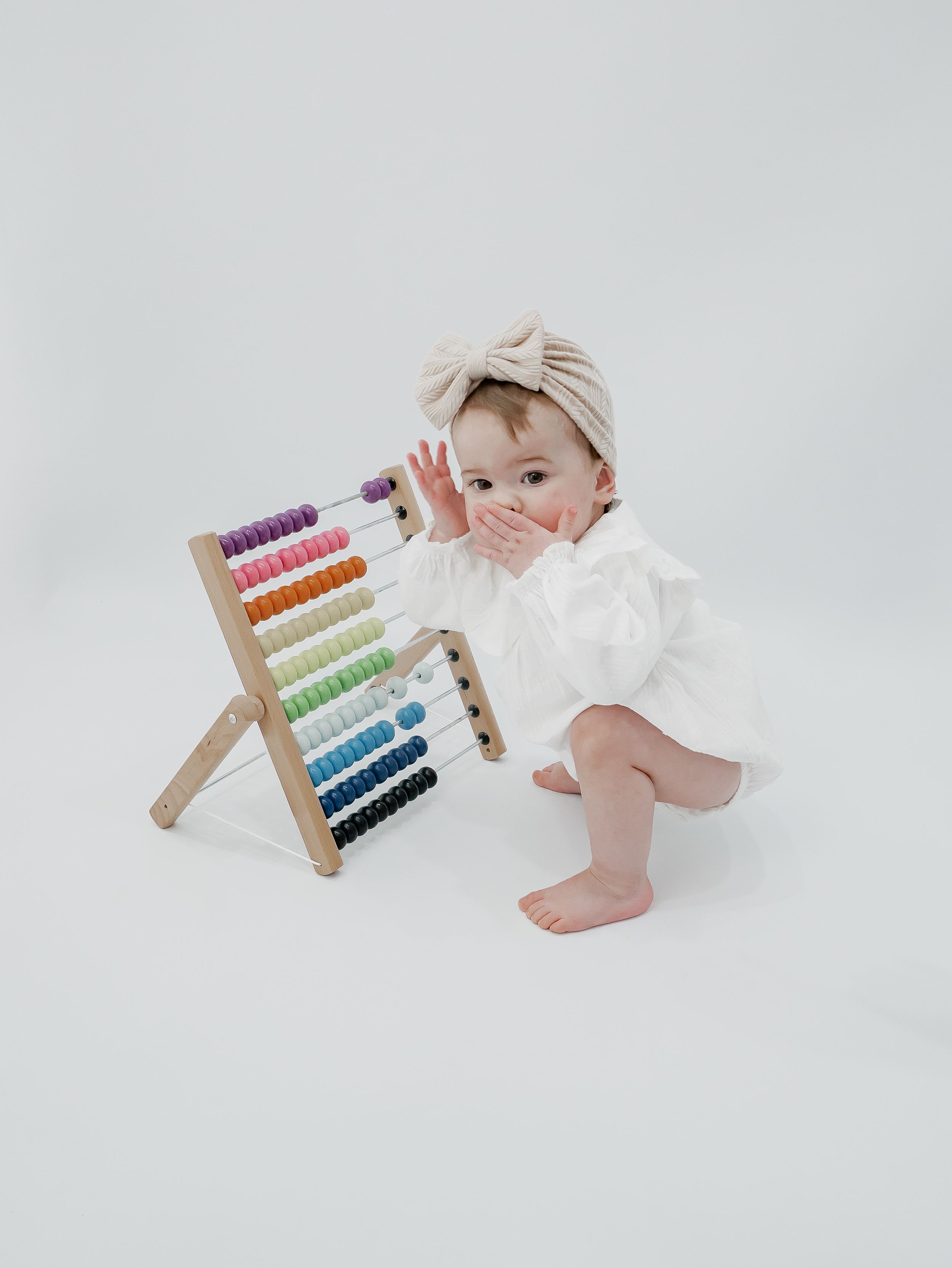 Baby in white outfit plays with colorful wooden abacus against white background.