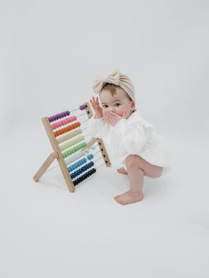 Baby in white outfit plays with colorful wooden abacus against white background.