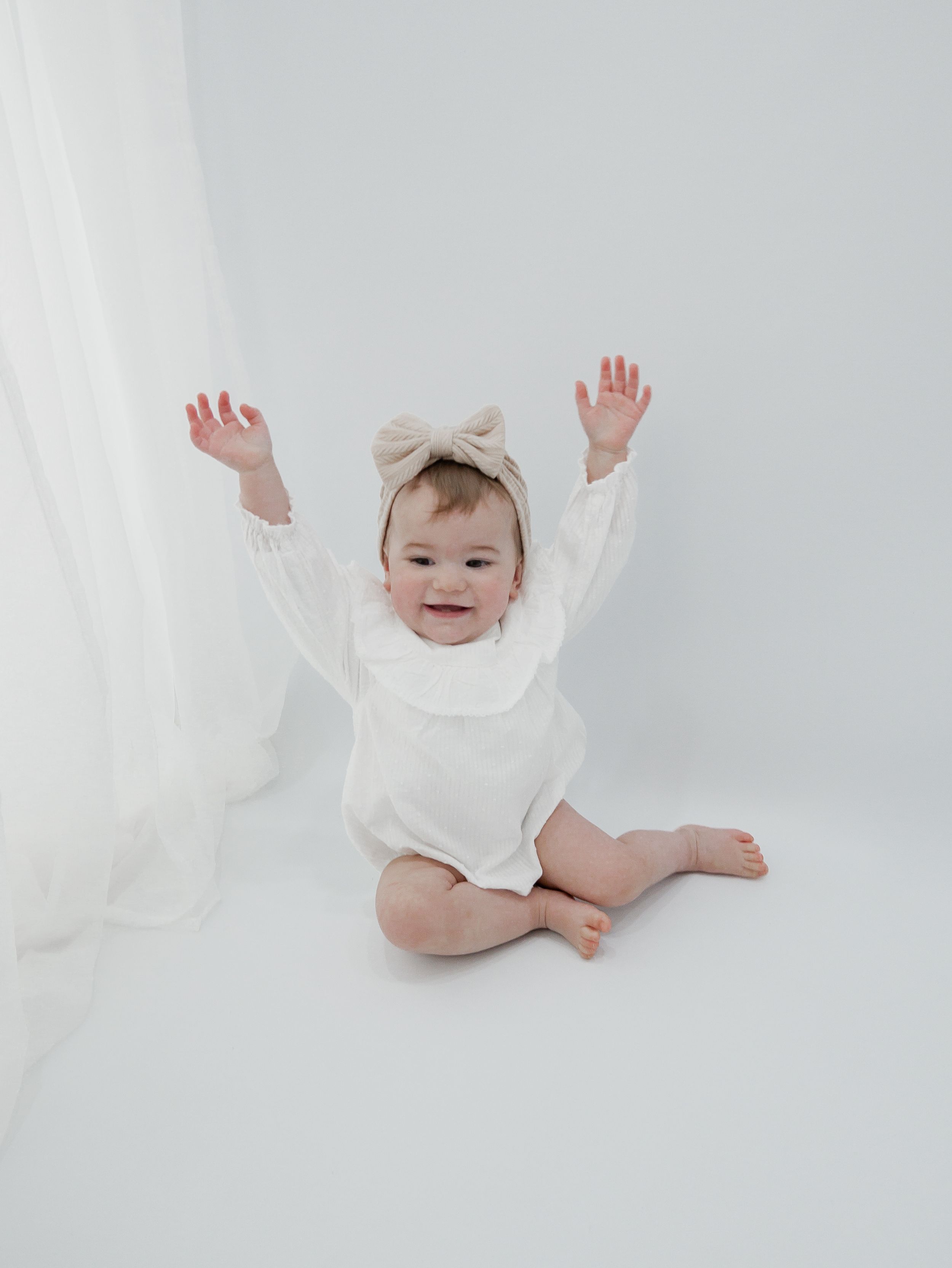Baby in white outfit raises arms joyfully while sitting on white floor.
