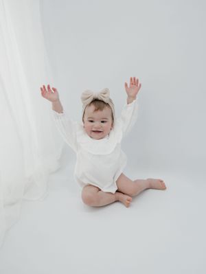 Baby in white outfit raises arms joyfully while sitting on white floor.