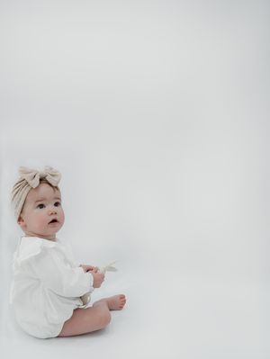 Baby wearing white clothing and a bow sits against minimal white background while looking off to the side.