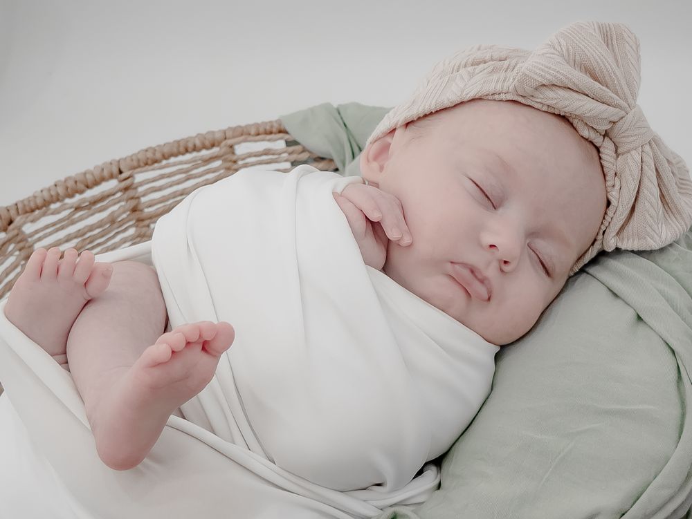 Peaceful sleeping baby wrapped in white with beige headwrap resting on soft bedding.