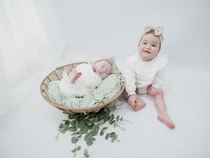 Two babies in white outfits pose in minimalist studio setting with wicker basket and greenery.