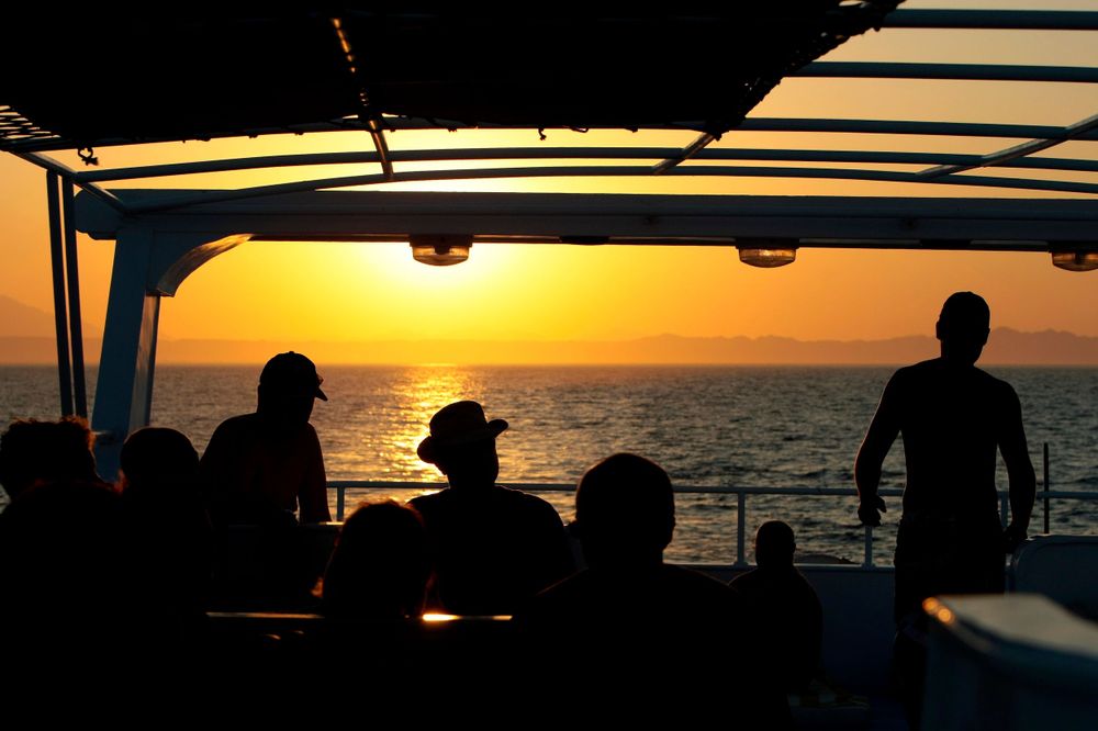 Silhouettes gather on a boat deck during a vibrant orange sunset over the ocean.