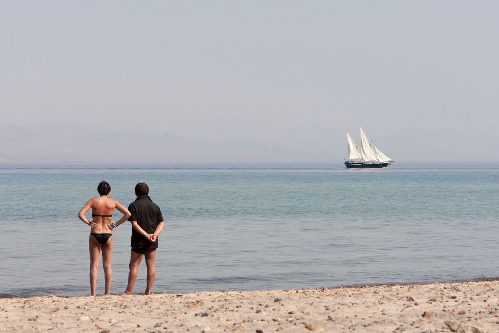 Two figures stand on a beach watching a sailboat glide across the distant horizon.
