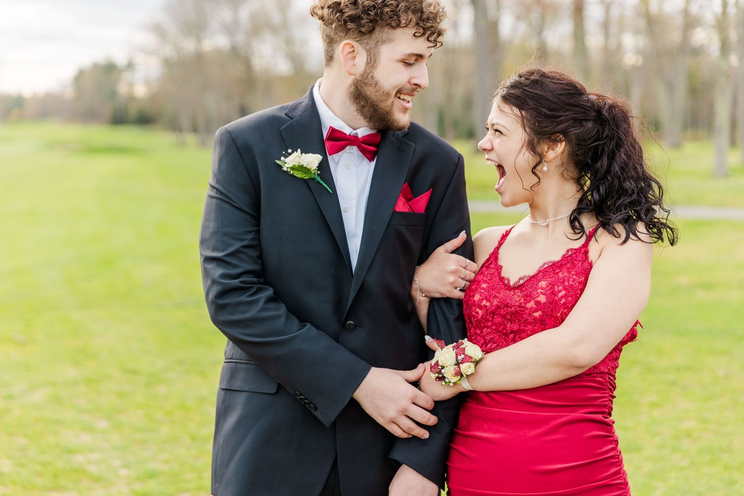 Formal couple in red dress and black tuxedo share a joyful moment together in an outdoor spring setting.
