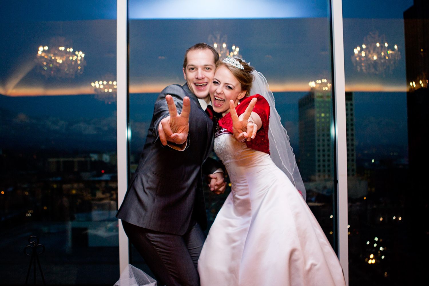 A joyful wedding couple points at the camera while posing by a window with city lights in the background.