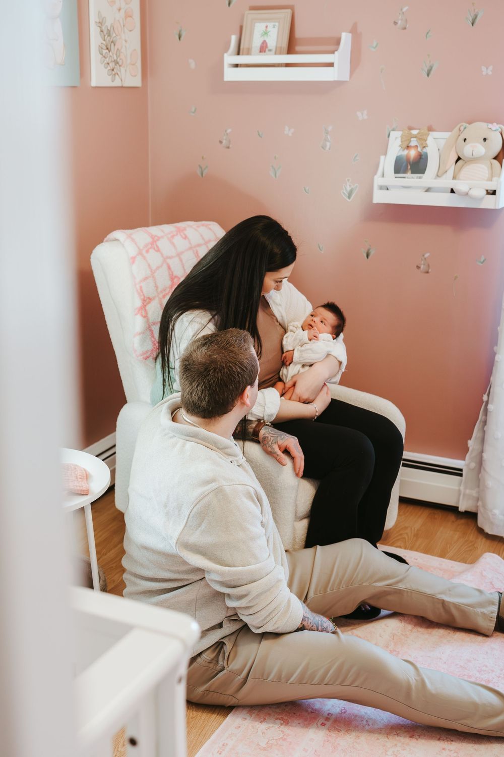 Parents bonding with newborn baby in pink nursery, mother holding infant in white glider chair.